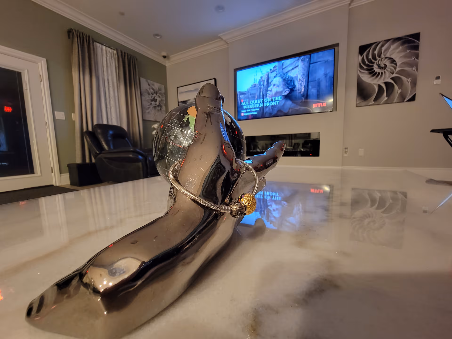 A modern living room with a shiny metallic hand sculpture holding a globe on a marble table in the foreground. In the background, there is a wall-mounted TV displaying a scene from a Netflix show titled 'All Quiet on the Western Front,' two black leather chairs, curtains, and framed artwork on the walls.
