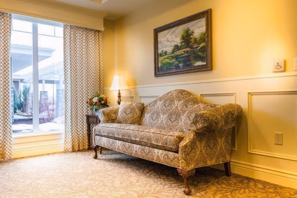 A cozy living room area with a patterned upholstered sofa, a side table with a lamp and a flower arrangement, a large window with patterned curtains letting in natural light, and a framed landscape painting on the wall.