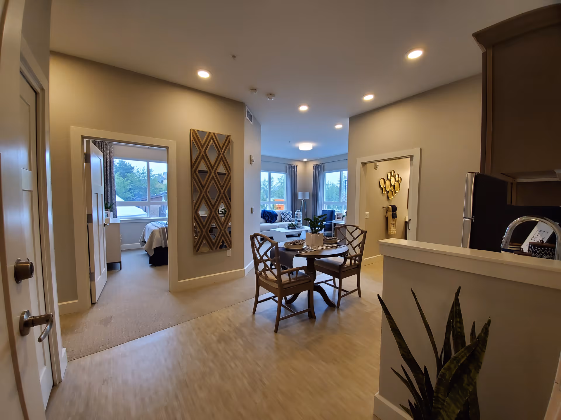 Interior view of a senior living apartment showing a small dining area with a round table and four chairs, a living room with a sofa and large windows in the background, and a bedroom visible through an open door on the left. The walls are painted light gray, and there is modern decor including a geometric wall mirror and a plant near the kitchen counter on the right.