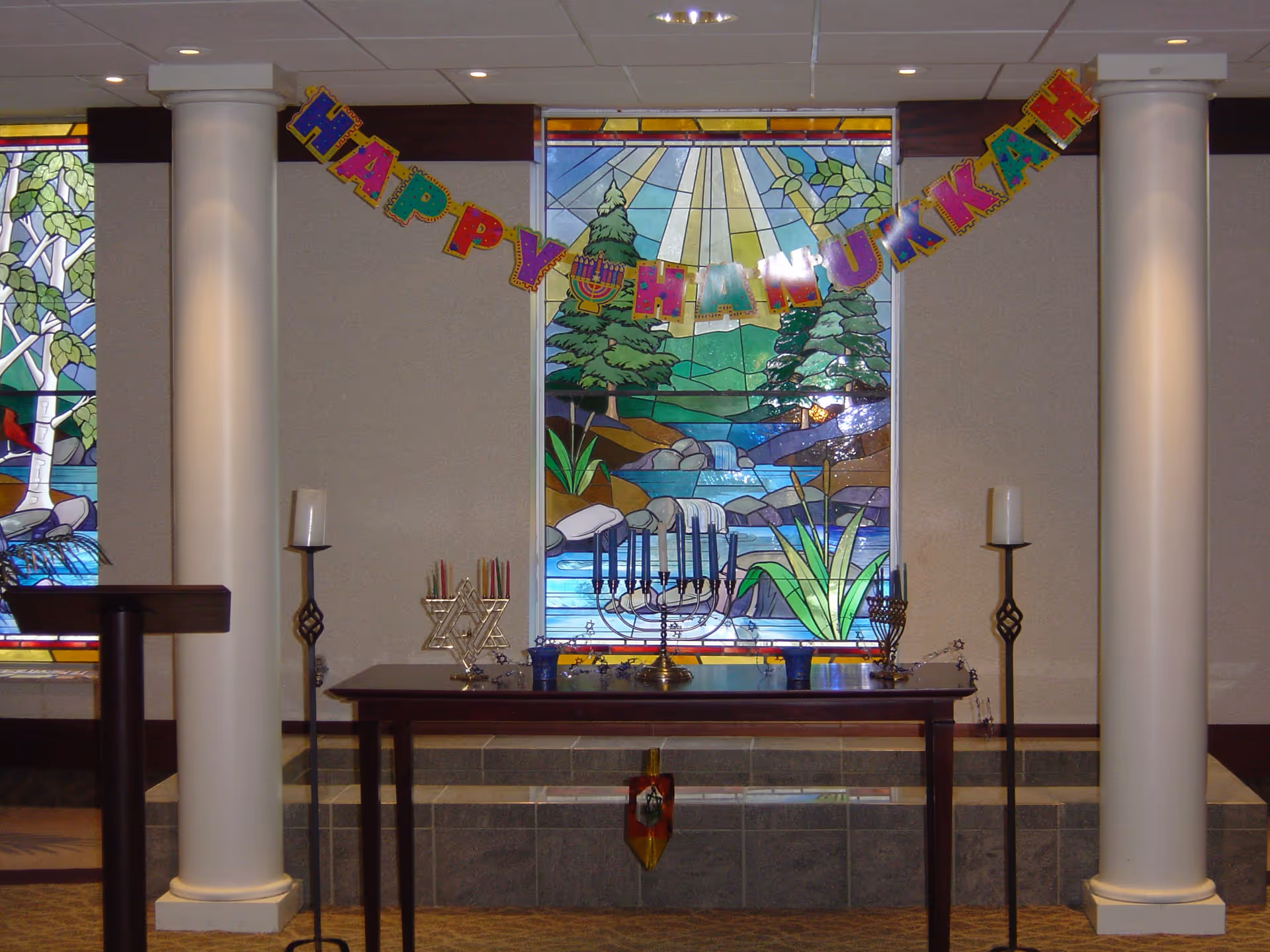 Interior view of a room with two white columns and a table in front of a stained glass window depicting a nature scene with trees, rocks, and a waterfall. On the table are three menorahs and some candles. Above the table hangs a colorful banner that reads 'HAPPY HANUKKAH'.