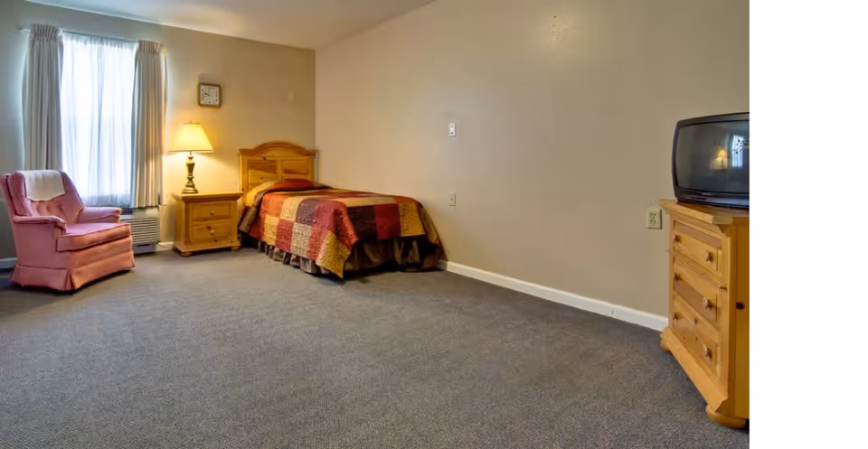 A simple bedroom with a single bed covered in a patchwork quilt, a wooden nightstand with a lamp, a pink upholstered armchair near a window with curtains, and a wooden dresser with an old-style TV on top. The walls are plain and the floor is carpeted.