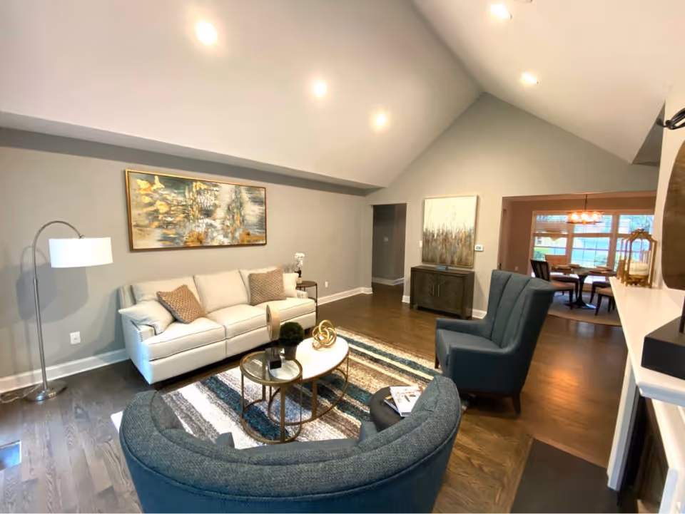 A modern living room with a white sofa, two blue armchairs, a round coffee table with decorative items, a floor lamp, and abstract artwork on the walls. The room has hardwood floors and recessed ceiling lights. In the background, a dining area with a table and chairs is visible through an open doorway.