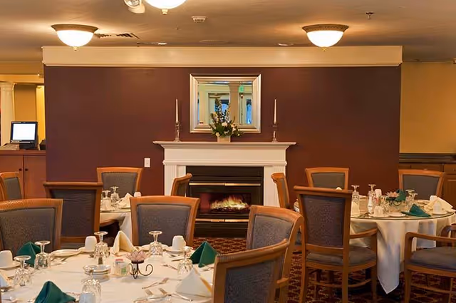 Dining room with round tables set with white tablecloths, green and white folded napkins, glassware, and silverware. Chairs with wooden frames and upholstered seats surround the tables. A fireplace with a white mantel and a mirror above it is visible against a dark brown wall.