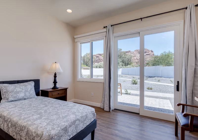 A furnished bedroom with bed, nightstand, lamp, and sliding glass door to a patio.