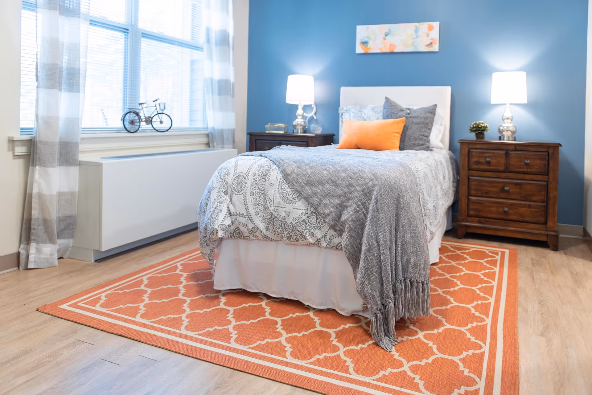 A cozy bedroom with a single bed featuring patterned bedding, a gray throw blanket, and decorative pillows including an orange one. The bed is positioned on an orange patterned rug. There are two wooden nightstands on either side of the bed, each with a lamp and small decorative items. The wall behind the bed is painted blue and has a colorful abstract painting. A window with sheer striped curtains allows natural light into the room.