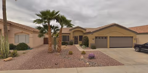 Front exterior view of a single-story residential building with a tiled roof, two palm trees, desert landscaping with rocks and small plants, a driveway leading to a two-car garage, and a cloudy sky overhead.