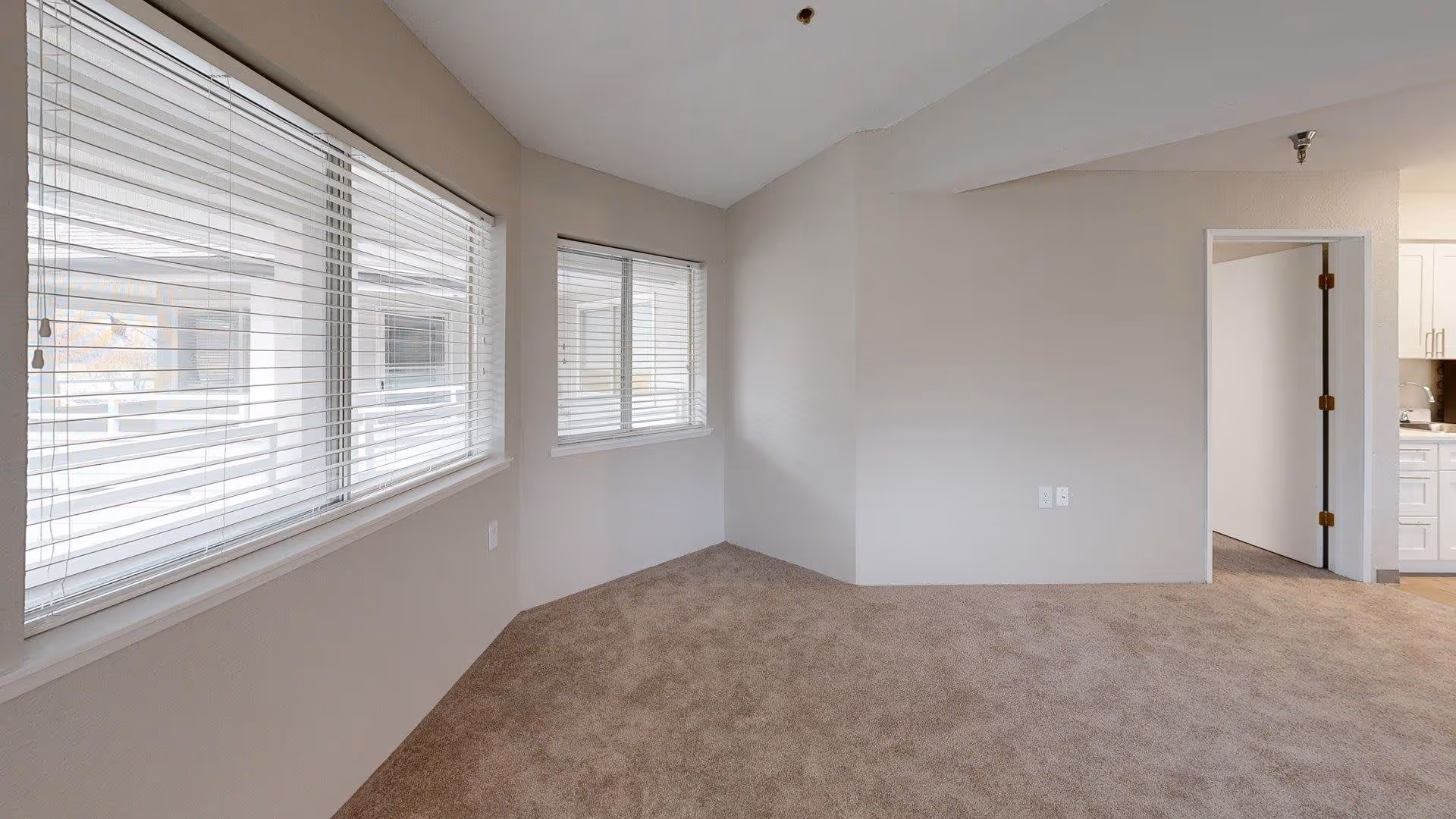 Empty interior room with beige carpet and light-colored walls. Two large windows with white blinds are on the left wall, allowing natural light to enter. An open doorway on the right leads to another room with white cabinetry visible.