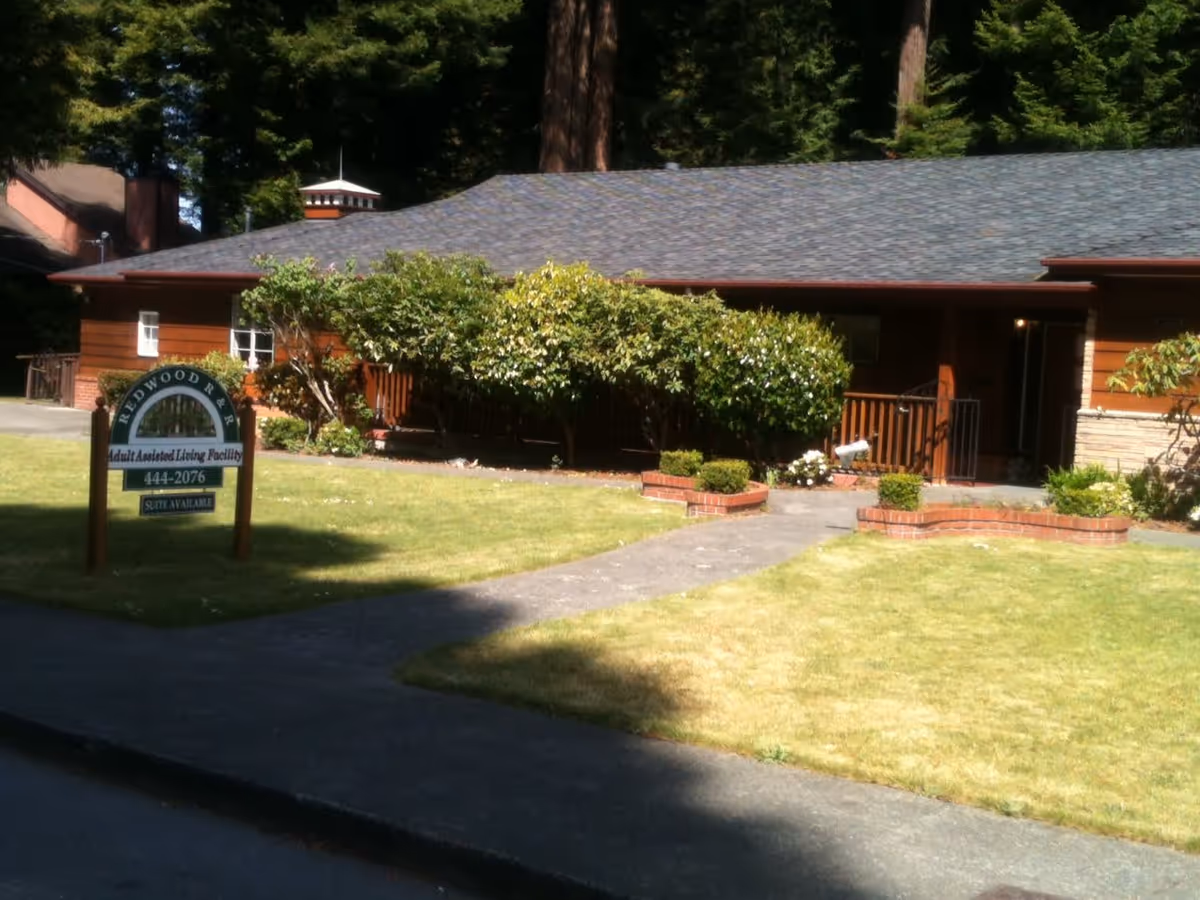 Single-story brown assisted living building with a front lawn, walkway, shrubs, and a 'Redwood R & R' sign near the sidewalk.