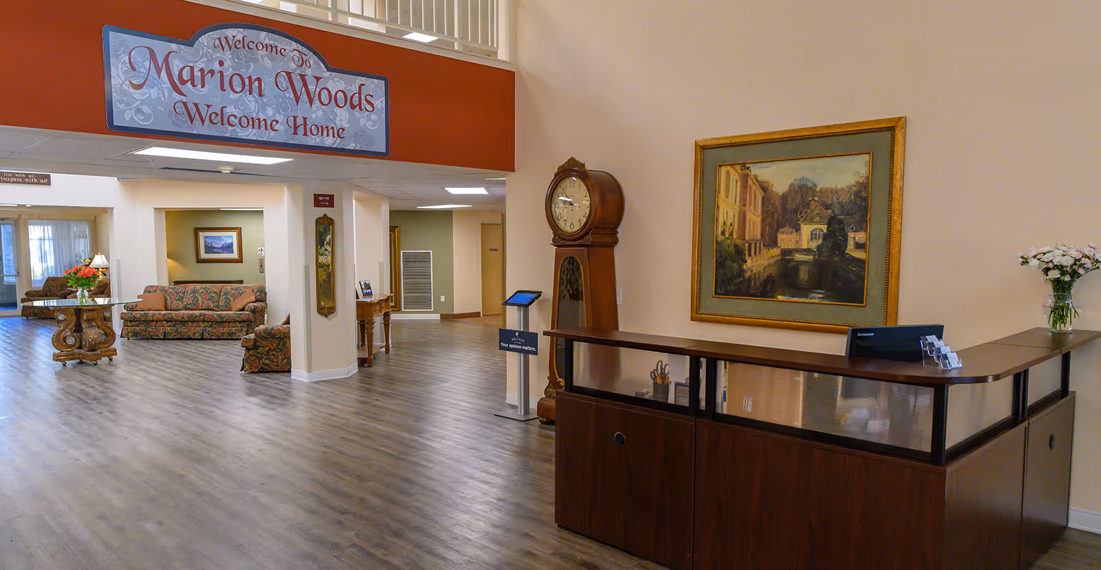 Interior view of the Marion Woods senior living facility entrance area featuring a reception desk with a vase of flowers, a large grandfather clock, a framed painting on the wall, and a seating area with floral-patterned couches in the background. A welcoming sign above reads 'Welcome to Marion Woods Welcome Home.'