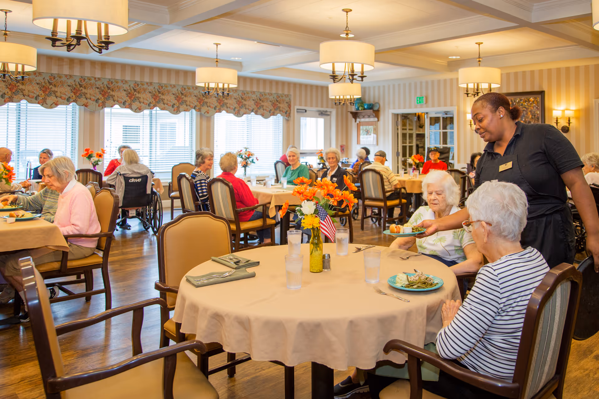A dining room in an assisted living facility with elderly residents seated at tables covered with beige tablecloths. A staff member is serving food to two elderly women at one table. The room is well-lit with large windows, floral curtains, and multiple ceiling lights. Tables have flower vases with orange and yellow flowers and some have small American flags.