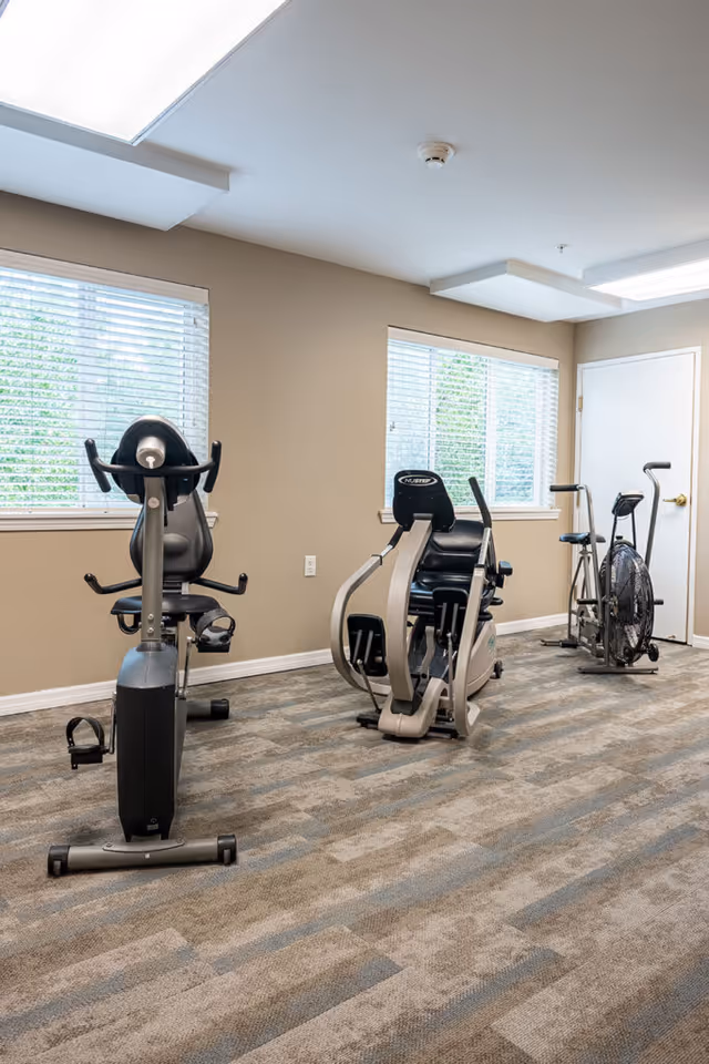 Small fitness room with stationary exercise bikes, carpeted floor, and windows on beige walls.