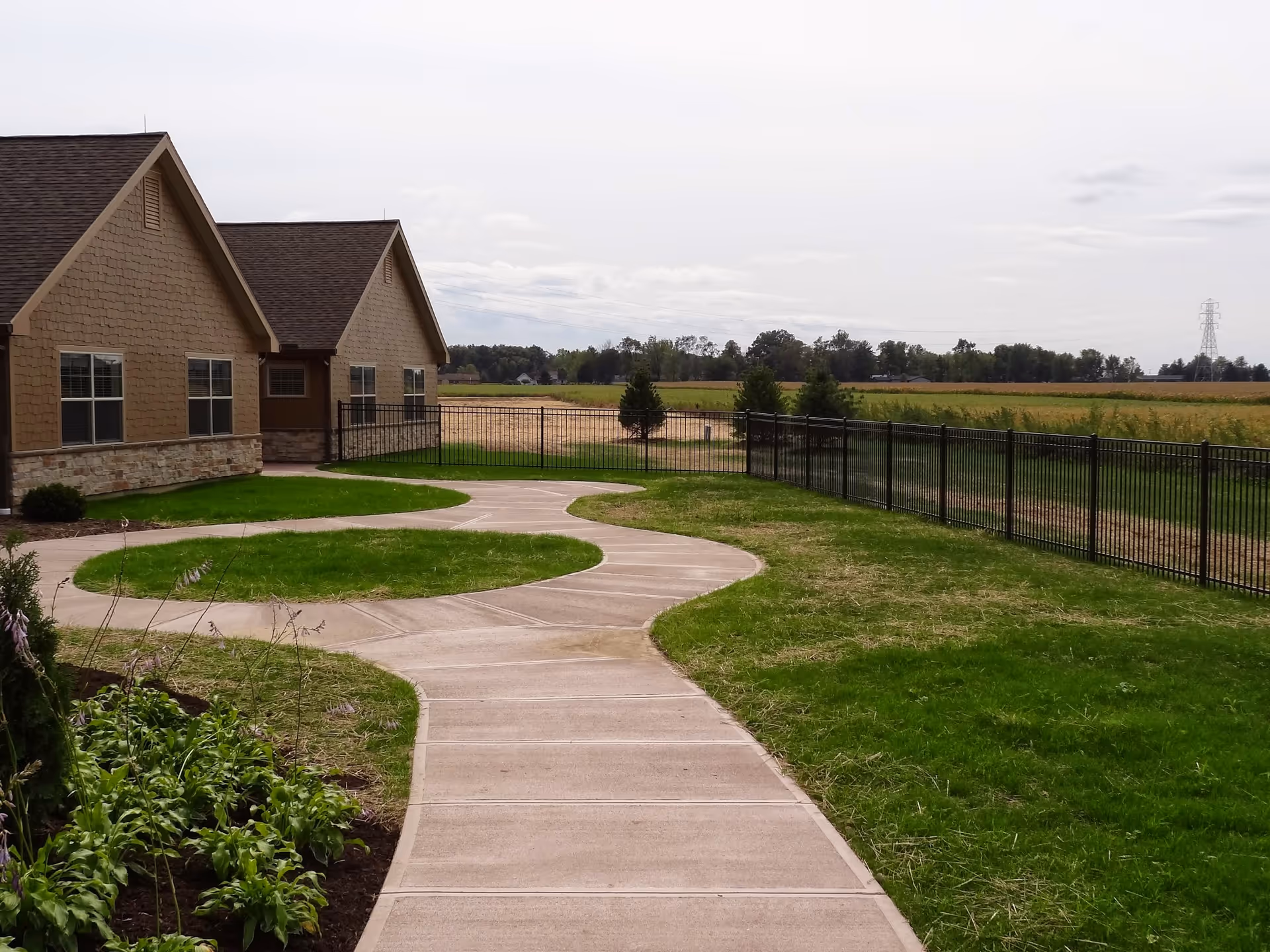 Curved concrete walkway leading to a beige building with multiple windows, surrounded by green grass and a black metal fence, with open fields and trees in the background under a cloudy sky.