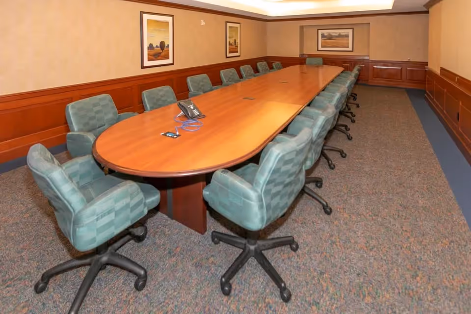 A long wooden conference table surrounded by green cushioned office chairs on wheels in a carpeted room with wood paneling on the walls and framed landscape paintings.