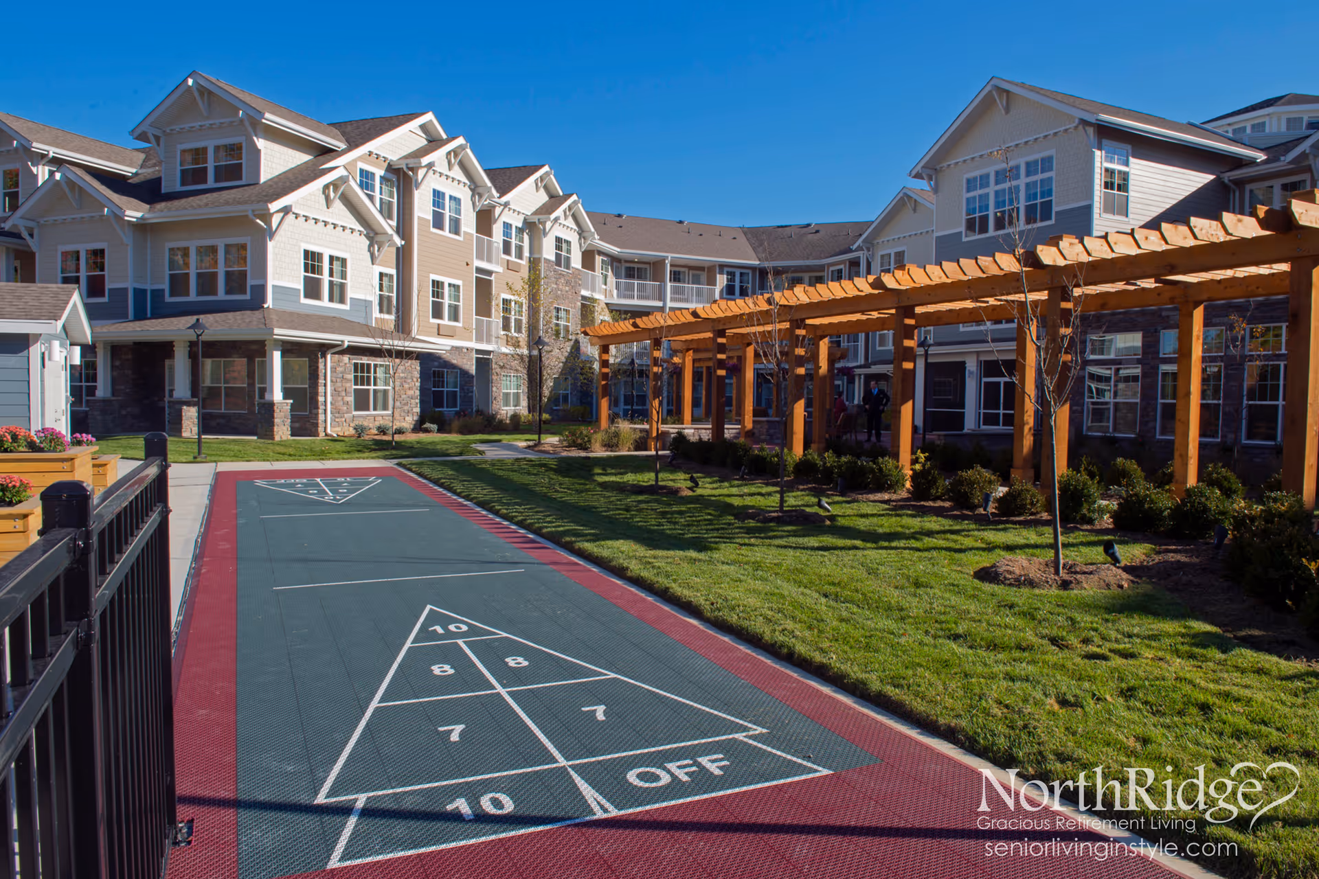 Outdoor courtyard area of Northridge Gracious Retirement Living featuring a shuffleboard court, green lawn, wooden pergola, and a multi-story residential building with beige and stone exterior under a clear blue sky.