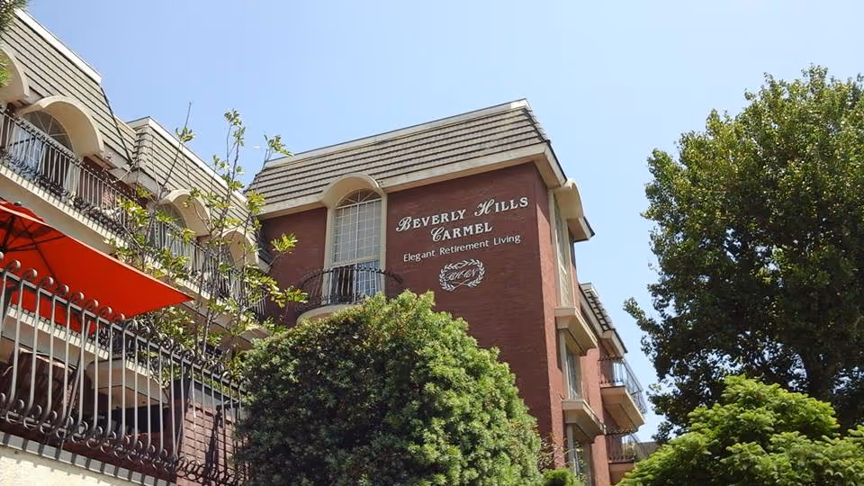 Exterior view of a multi-story brick building with balconies and black wrought iron railings. The building has a sign that reads 'Beverly Hills Carmel Elegant Retirement Living'. There are trees and greenery in the foreground under a clear blue sky.