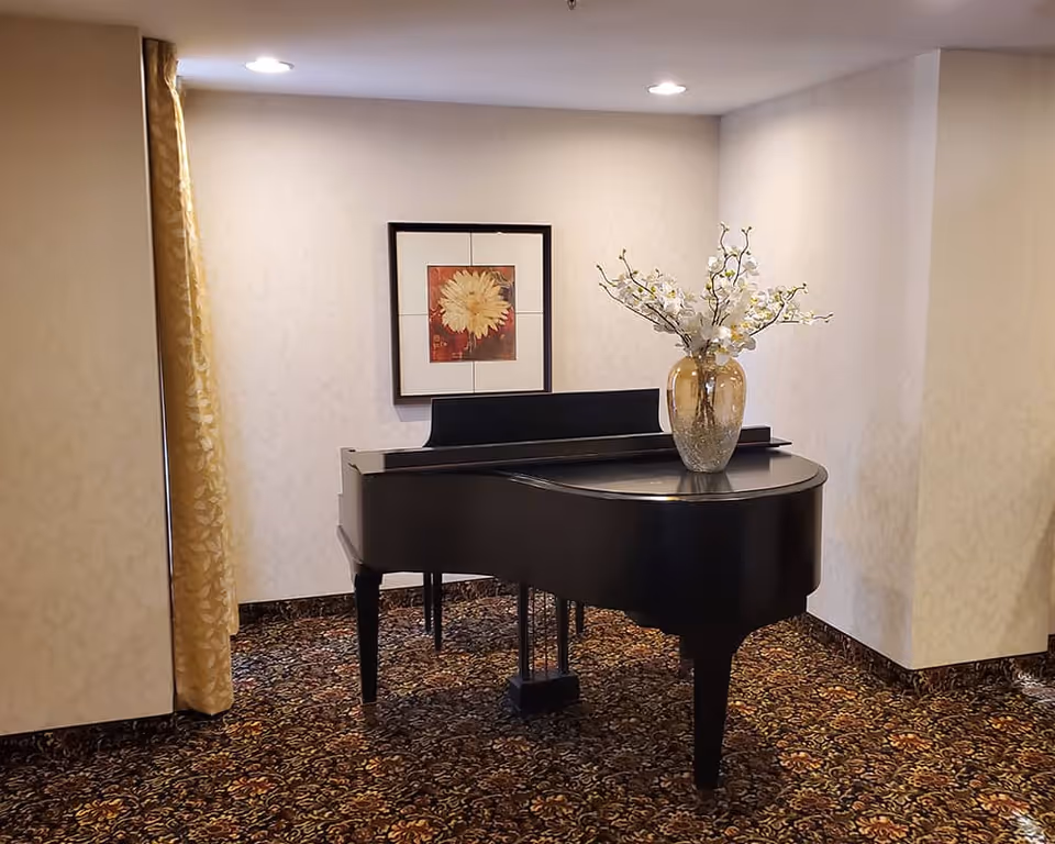 A black grand piano with a vase of white flowers on top is placed in a carpeted corner of a room with cream-colored walls and a framed floral picture hanging above the piano.