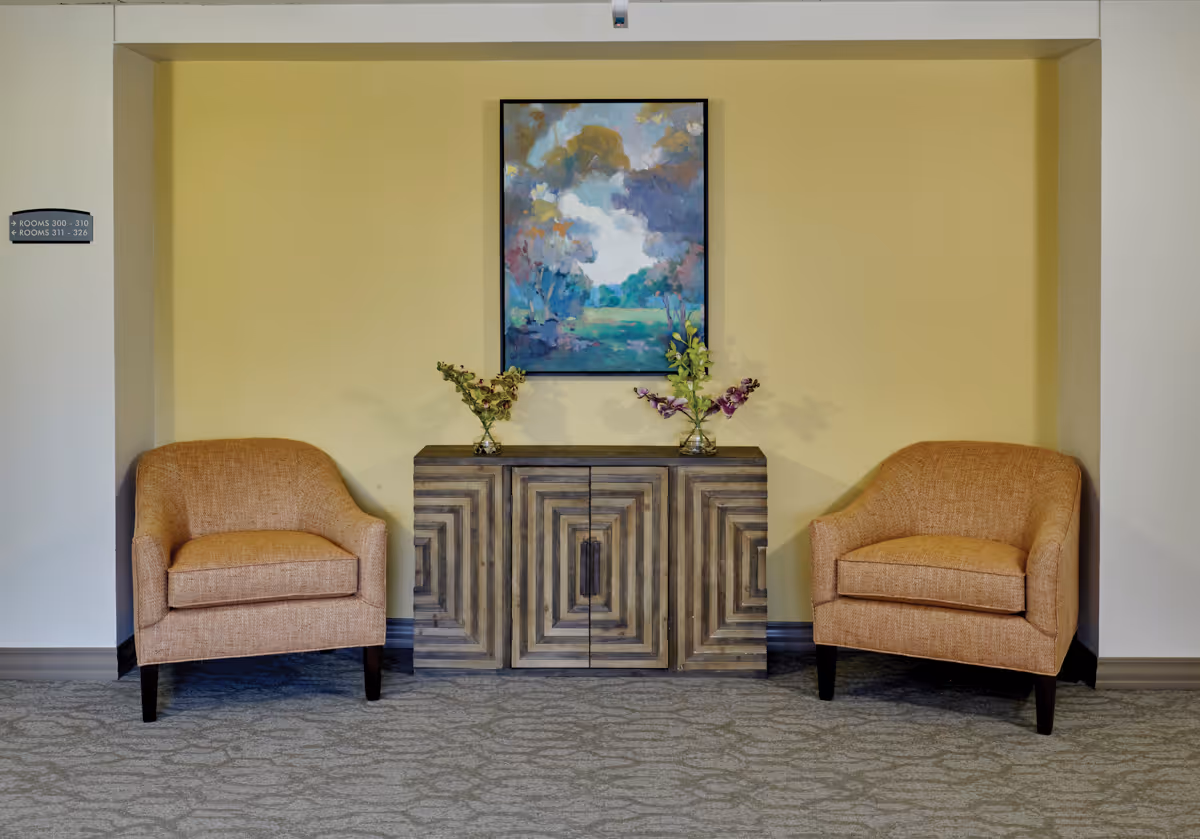 Two orange upholstered armchairs placed on either side of a wooden cabinet with a geometric pattern. On top of the cabinet are two small vases with flowers. Above the cabinet hangs a colorful landscape painting. The walls are painted light yellow and there is a sign on the left wall indicating room numbers.