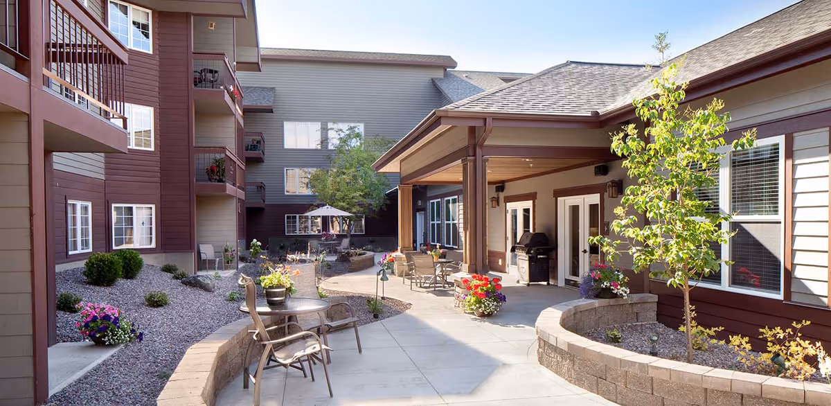 Courtyard with patio chairs, flowering planters, and a covered patio area surrounded by a multi-story senior living building.