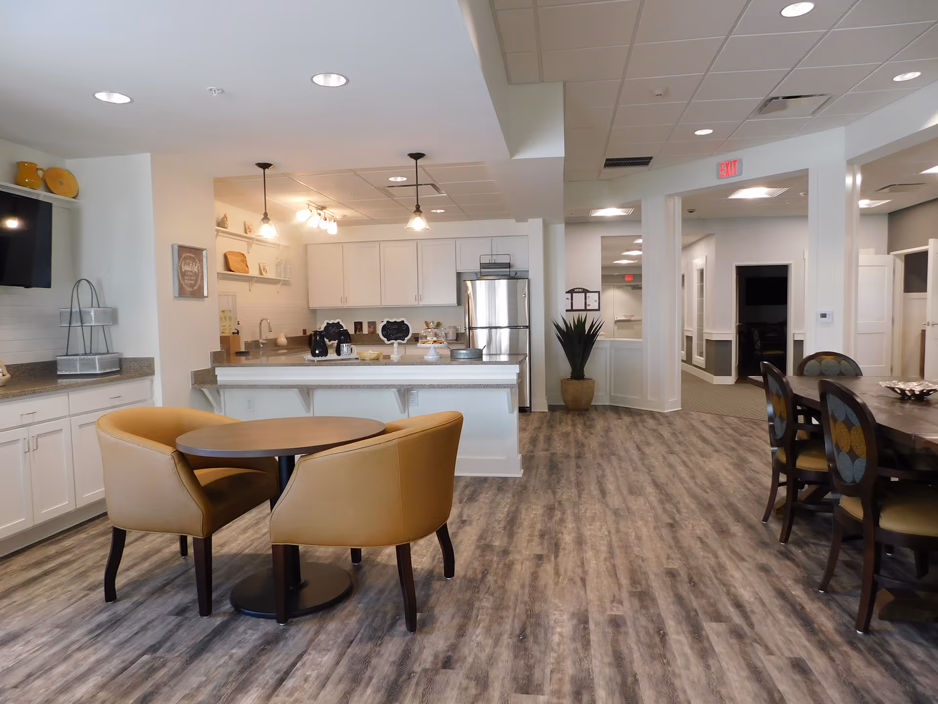 Communal dining area with round tables and chairs in front of an open kitchen and island with a stainless refrigerator.