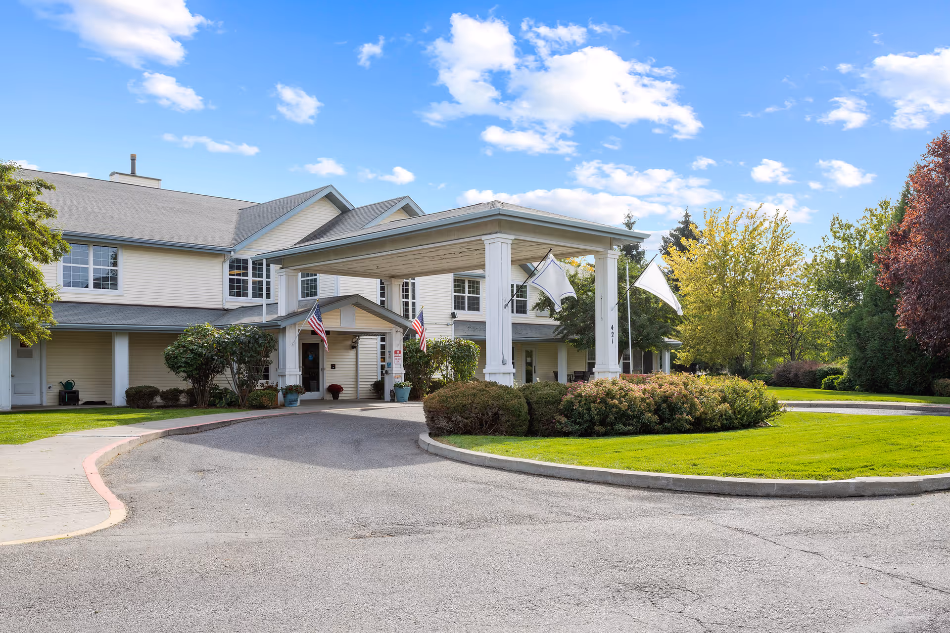 Front entrance of a two-story senior living building with a covered porte-cochère, flags, and landscaped driveway.