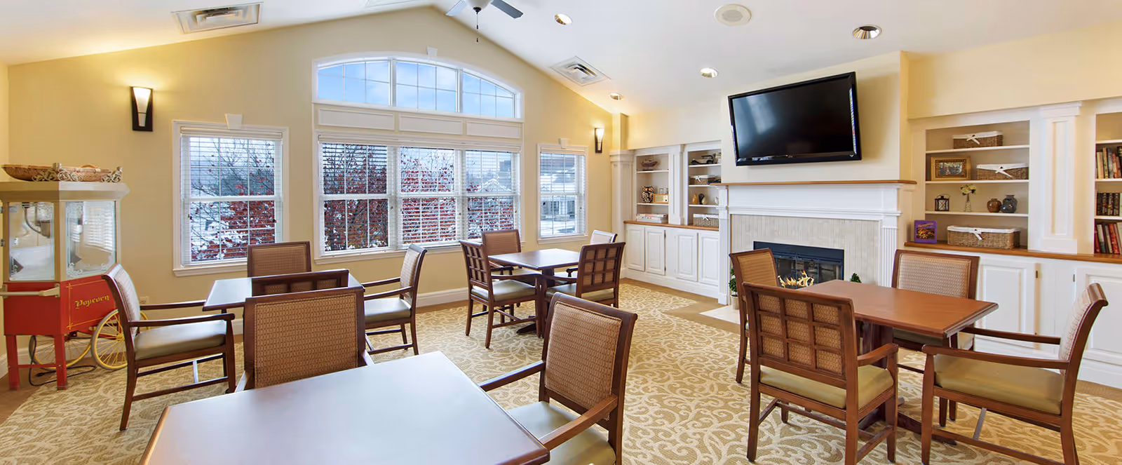 A bright and cozy common area with multiple wooden tables and chairs arranged on a patterned carpet. Large windows with white blinds allow natural light to fill the room. There is a fireplace with a mounted flat-screen TV above it, and built-in white shelves on either side holding decorative items and books. A popcorn machine is visible in the corner near the windows.