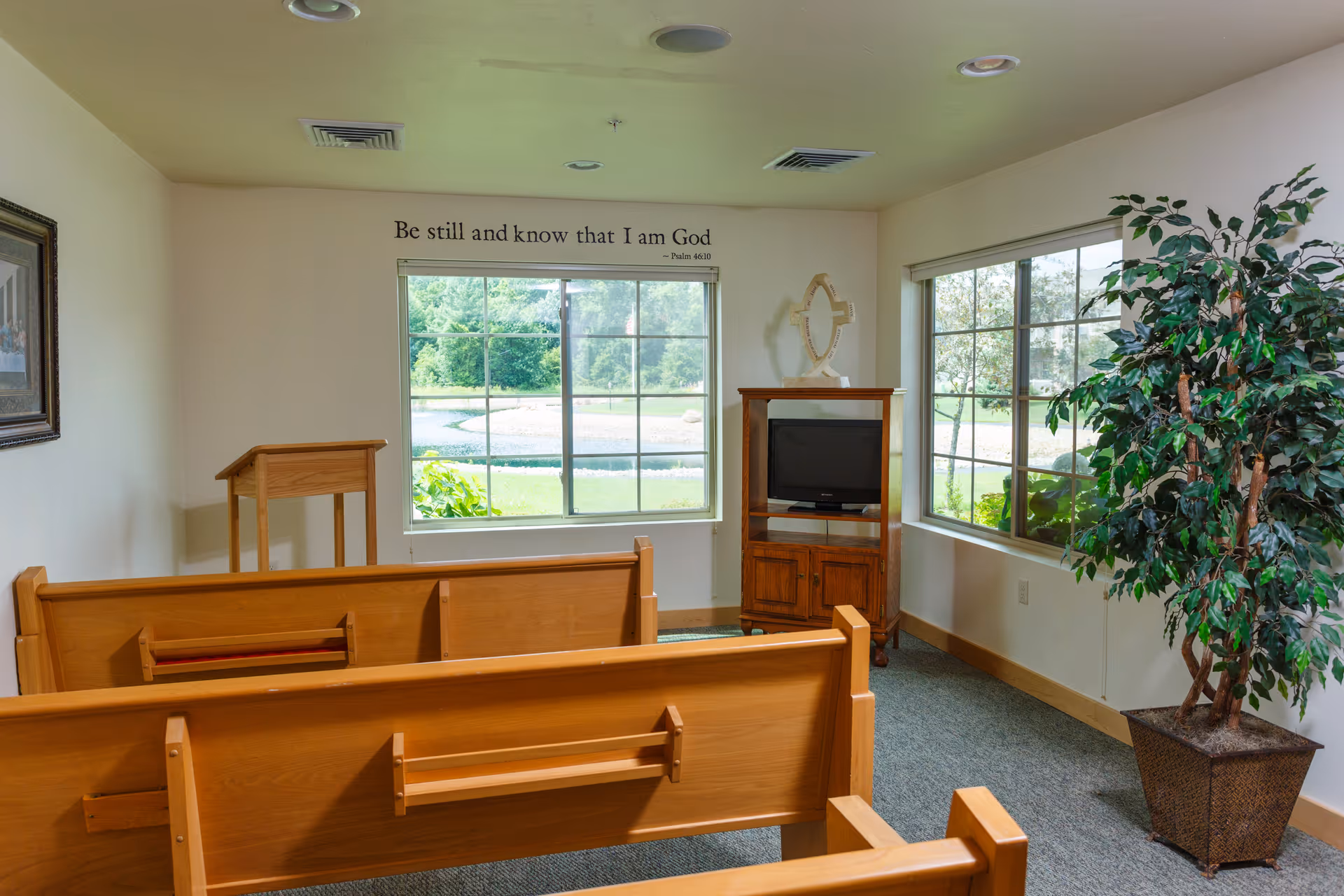 A small chapel room with wooden pews facing a wooden podium and a TV cabinet. Two large windows show a green outdoor landscape. A decorative cross is on top of the TV cabinet. On the wall above one window is a religious quote: 'Be still and know that I am God - Psalm 46:10'. A potted plant is in the corner near the other window.