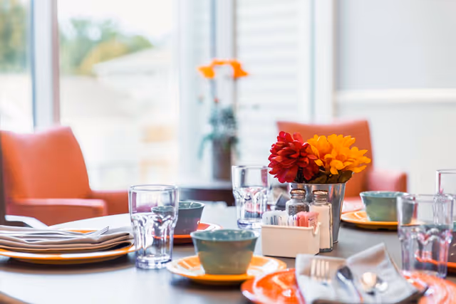 A dining table set with colorful plates, bowls, glasses and a small floral centerpiece in a bright dining area.