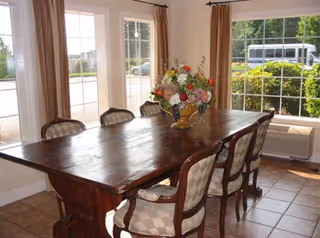 Sunlit dining room featuring a long wooden table surrounded by upholstered chairs and a floral centerpiece in front of large windows.
