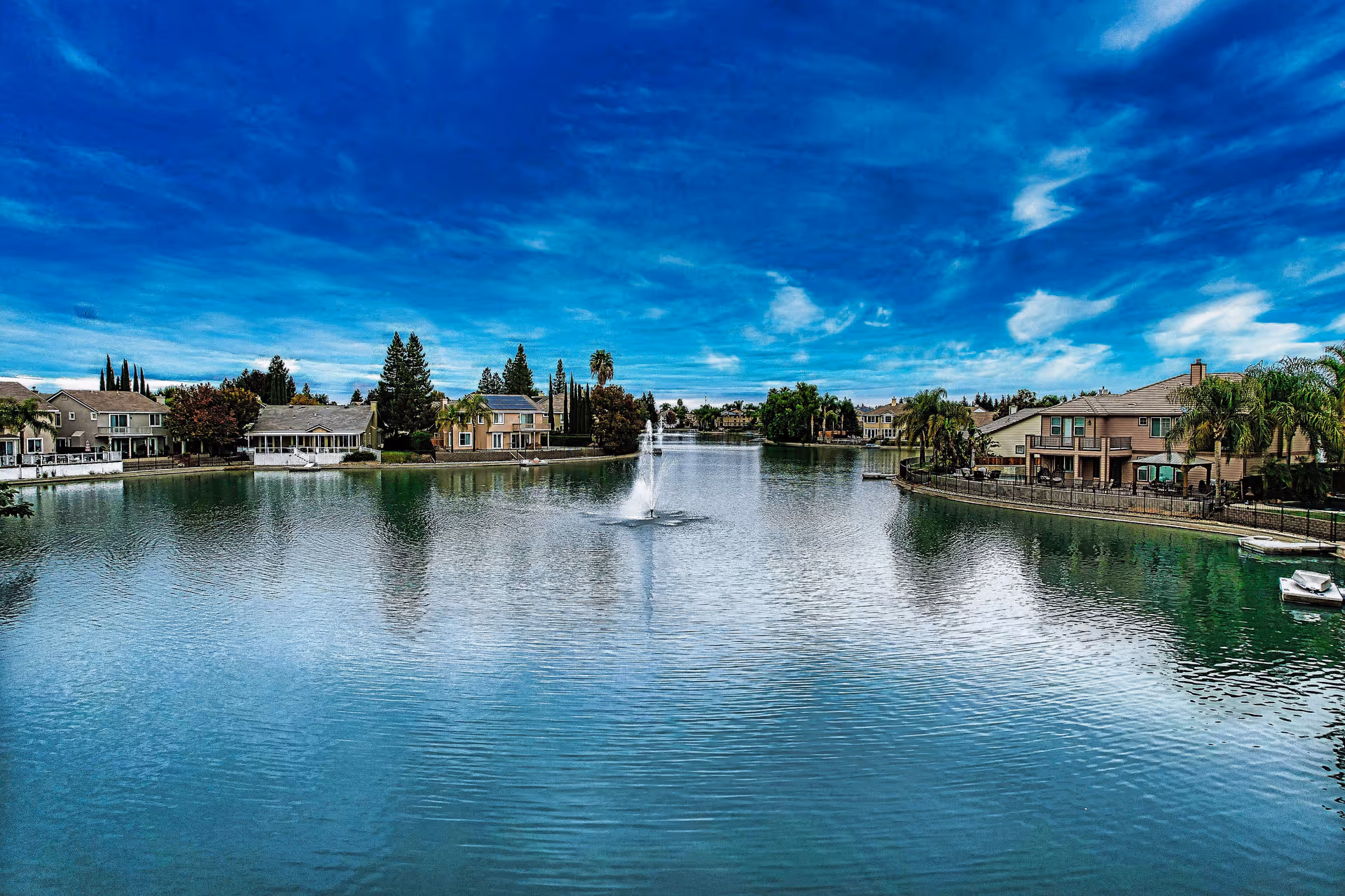 A calm residential lake with a central fountain and waterfront houses under a blue sky.