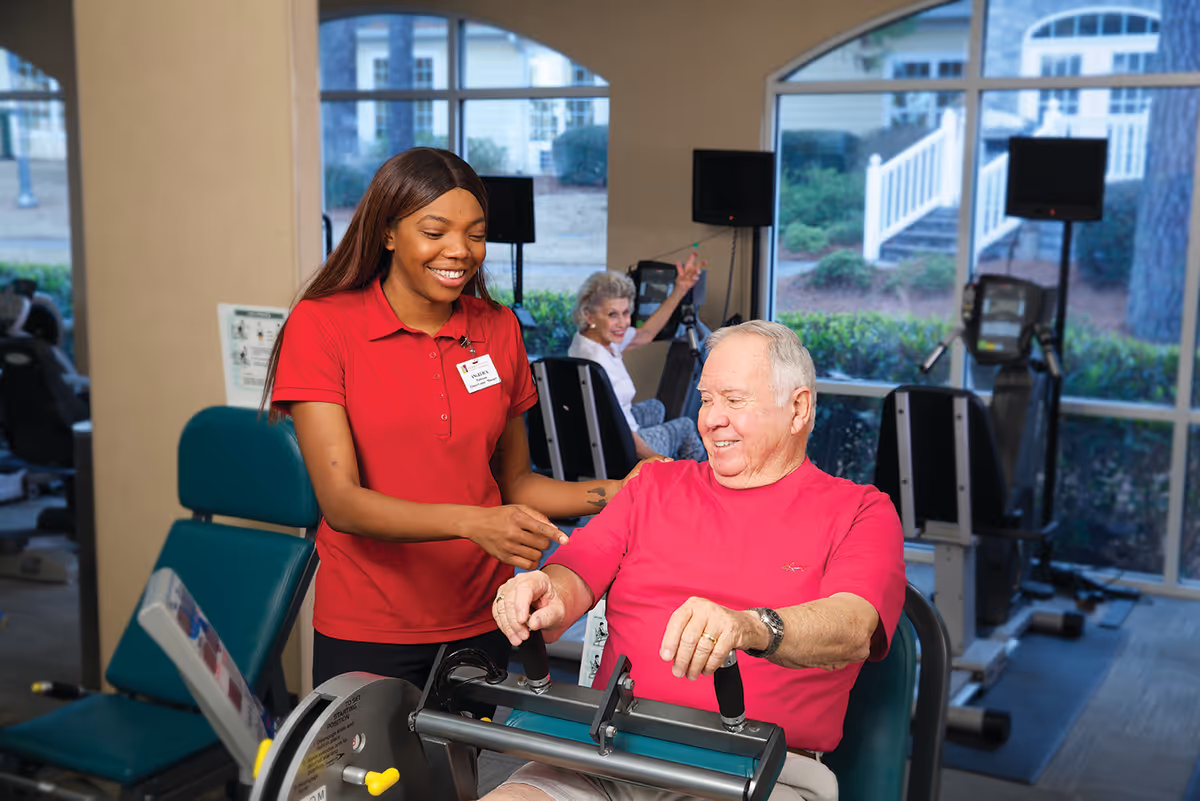 A smiling female staff member in a red polo shirt assists an elderly man using a seated exercise machine in a fitness room with large windows. Another elderly woman is visible in the background using exercise equipment.