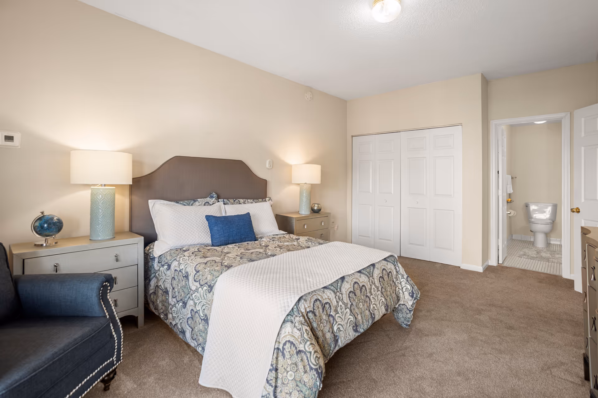 A bedroom with a bed featuring a patterned comforter and multiple pillows, two bedside tables each with a lamp, a blue armchair, a closet with white double doors, and an open door leading to a bathroom with a visible toilet.