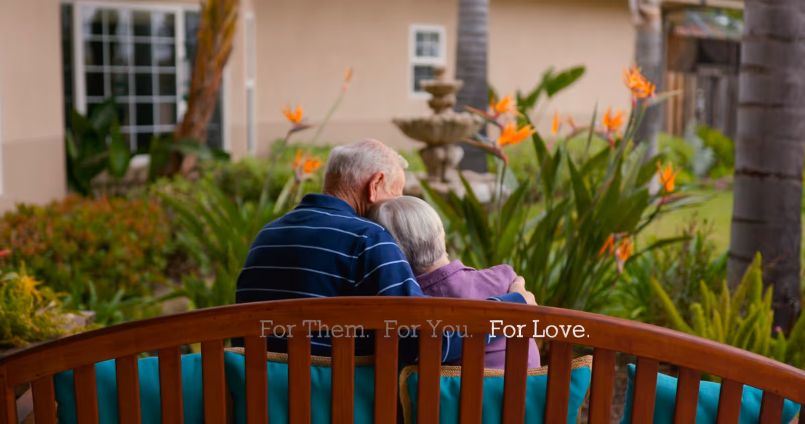 An elderly couple sitting closely together on a wooden bench in a garden area with lush greenery and orange flowers, with a building and windows in the background.