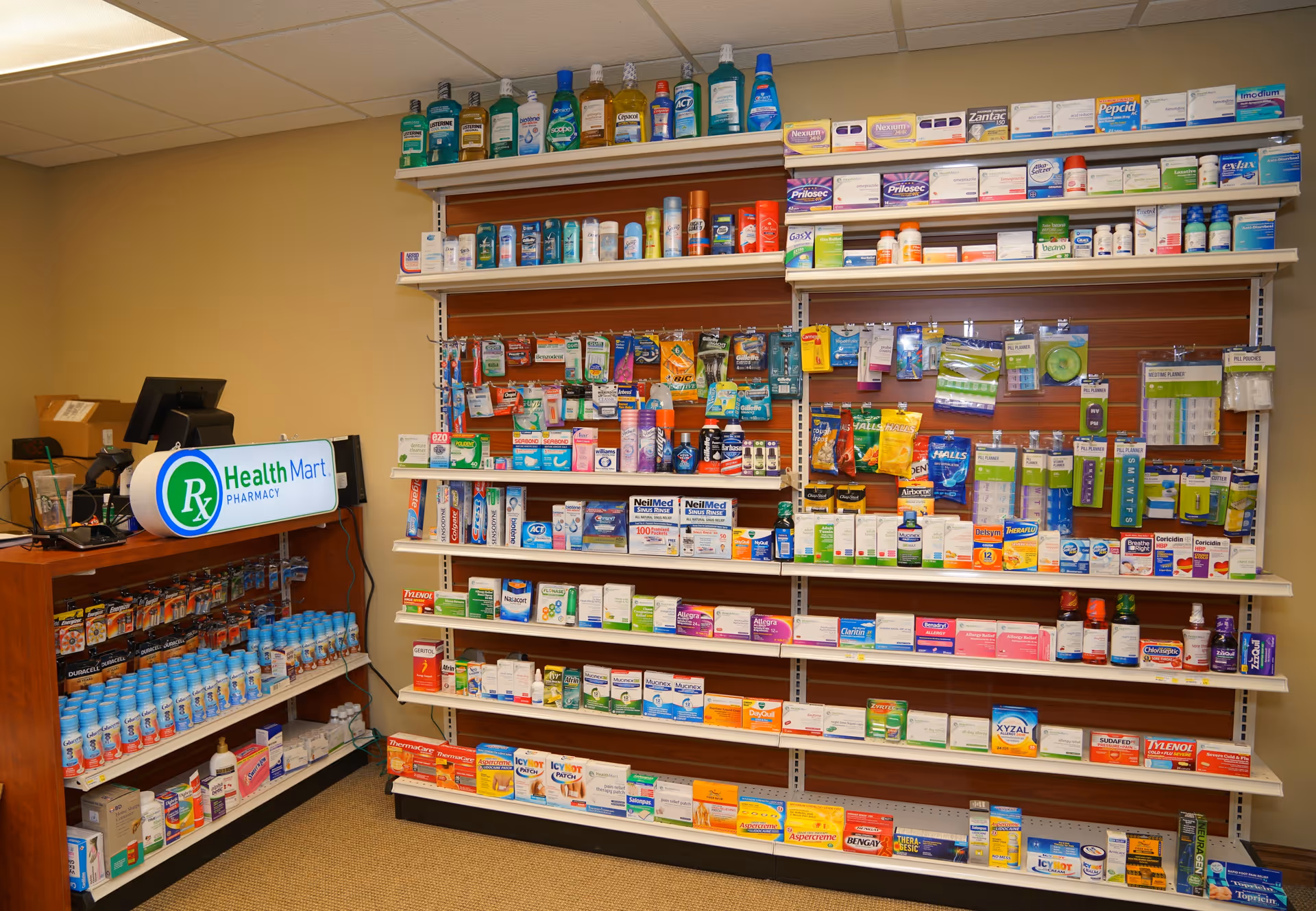 Interior view of a pharmacy area with shelves stocked with various over-the-counter medications, health products, and personal care items. A counter with a Health Mart Pharmacy sign is visible on the left side.