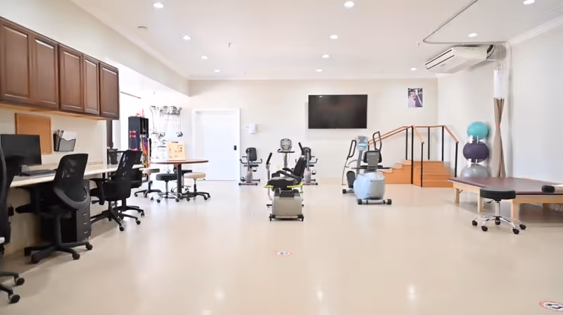 A spacious rehabilitation or therapy room with exercise equipment including stationary bikes and therapy balls. There are desks and chairs along the left wall, a large flat-screen TV mounted on the far wall, and a small set of stairs with a handrail on the right side. The room is well-lit with recessed ceiling lights and has a clean, minimalist design.