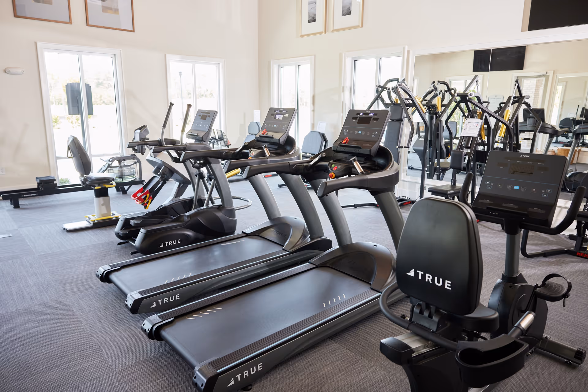 A bright fitness room with multiple exercise machines including treadmills, stationary bikes, and weight machines. The room has large windows allowing natural light to enter, and a large mirror on one wall reflecting the equipment.