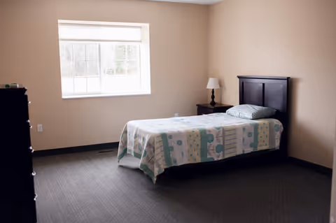 A simple bedroom with a single bed against the right wall, a nightstand with a lamp, a dresser, and a window letting in natural light.