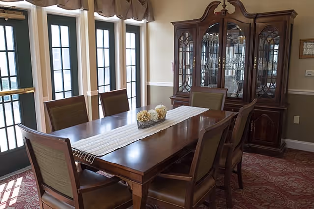A dining room with a wooden dining table surrounded by six chairs. The table has a white runner with a decorative centerpiece of flowers. Behind the table is a large wooden china cabinet with glass doors displaying dishes and glassware. The room has multiple tall windows with green frames and beige curtains, and a patterned red carpet on the floor.