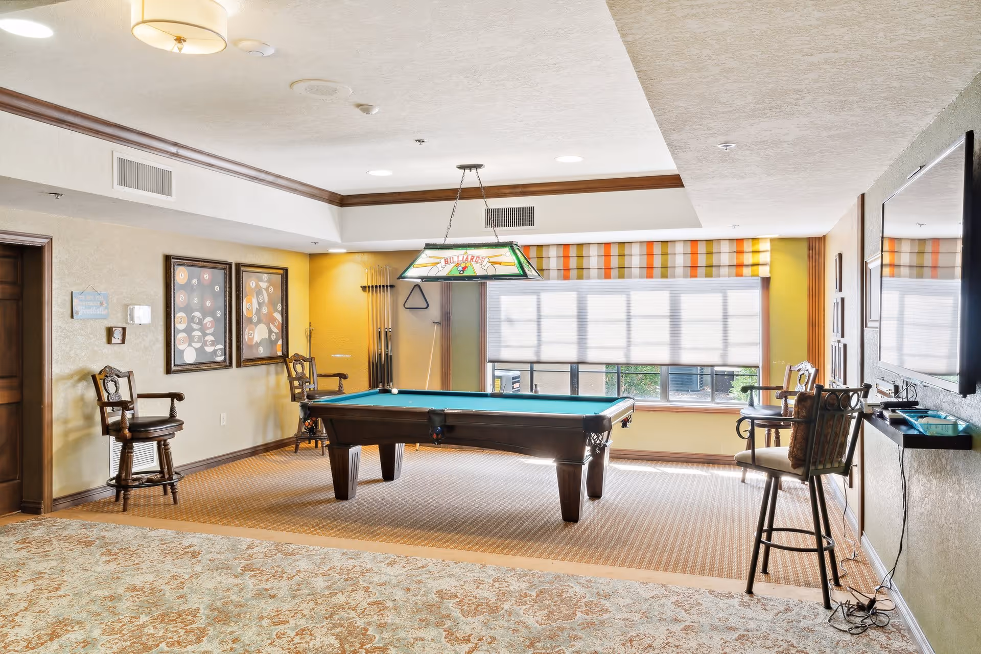 A well-lit recreational room featuring a pool table with a hanging billiards light above it. The room has a large window with striped valances and blinds, several wooden chairs with cushions, framed artwork on the walls, and a mounted flat-screen TV. The floor is carpeted with a patterned design near the pool table and a different carpet in the foreground.