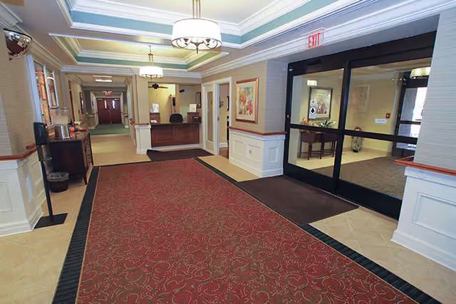 Interior view of a retirement facility lobby with a red patterned carpet, a reception desk in the background, framed artwork on the walls, and glass double doors on the right side.