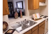 A kitchen area with a double stainless steel sink, wooden cabinets, a white electric stove with four burners, and a white countertop. Decorative items including a cookbook on a stand and two decorative bottles are placed on the counter. A pass-through window opens to a living area with a framed picture on the wall.