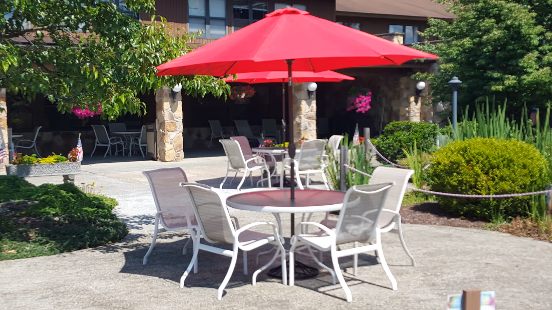 Outdoor patio area with round tables and white chairs under large red umbrellas, surrounded by greenery and bushes, with a building featuring stone pillars and windows in the background.