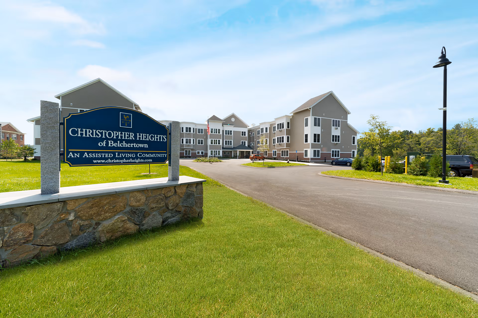 Exterior view of Christopher Heights of Belchertown, an assisted living community, showing a large building with multiple windows and a driveway. In the foreground, there is a stone sign with the community's name and website. The area is surrounded by green grass and trees under a partly cloudy sky.