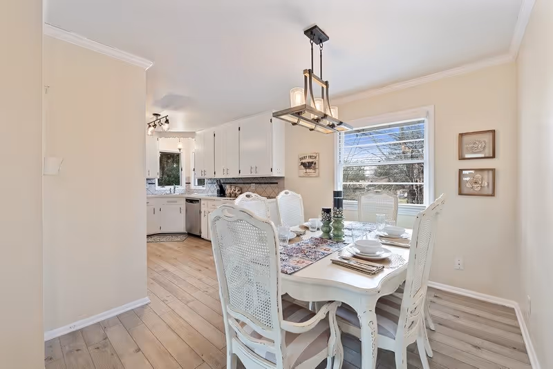 A bright dining area with a white wooden table set for six people, featuring white chairs with cane backs. The table has a decorative runner and candles in the center. In the background, there is a kitchen with white cabinets, a dishwasher, and a window letting in natural light. The walls are painted beige and the floor has light wood planks.