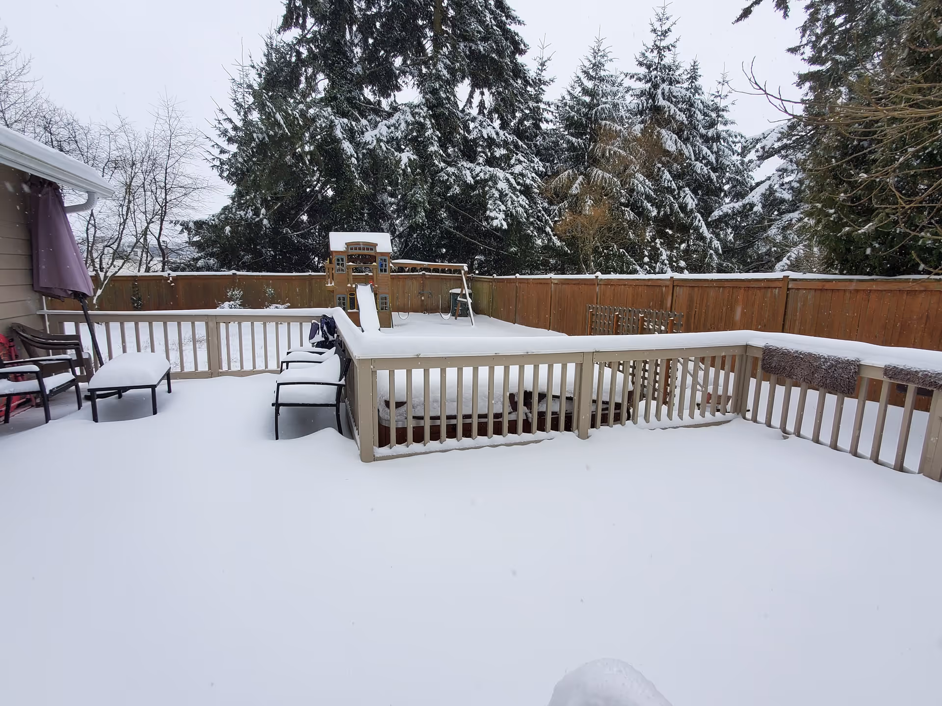 Snow-covered backyard deck with outdoor furniture including chairs and a table. A wooden play structure with a slide is visible in the background, surrounded by a wooden fence and snow-covered trees.