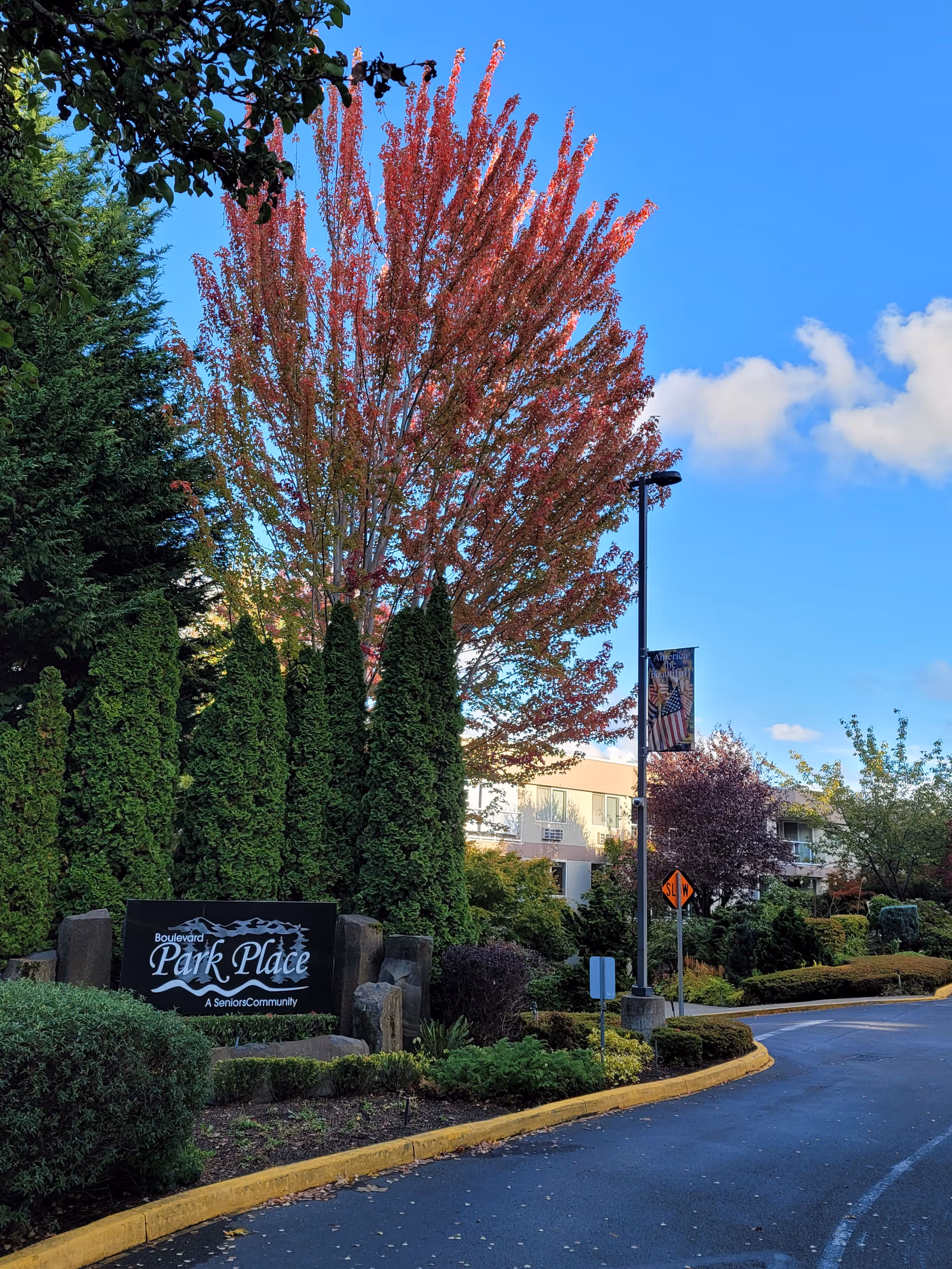 Entrance to Boulevard Park Place Active Retirement Living community with a landscaped area featuring tall green shrubs, a tree with red leaves, and a black sign displaying the community name. A curved driveway and a lamppost with a banner are also visible under a clear blue sky.