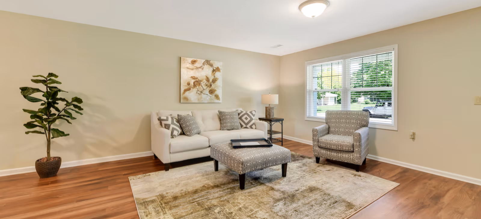 A bright living room with a beige sofa adorned with patterned cushions, a matching armchair, and a large ottoman on a textured area rug. There is a potted plant in the corner, a side table with a lamp, a framed artwork on the wall, and a window letting in natural light.