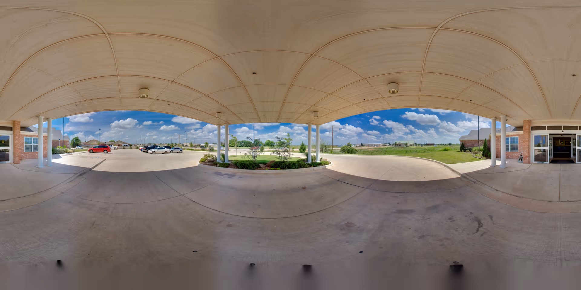 Covered entrance area of Plantation Village Nursing Center showing a wide concrete driveway with a few parked cars, green landscaping with bushes and trees, and a clear blue sky with scattered clouds.