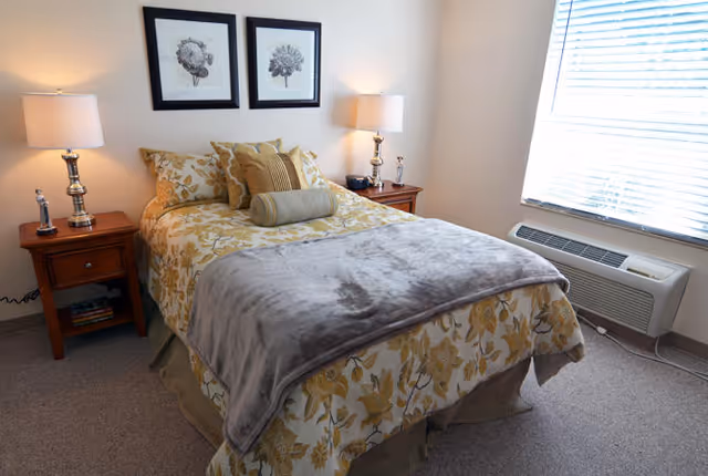Neatly made bed with floral bedding, two nightstands with lamps, and a window with blinds and an air conditioning unit.