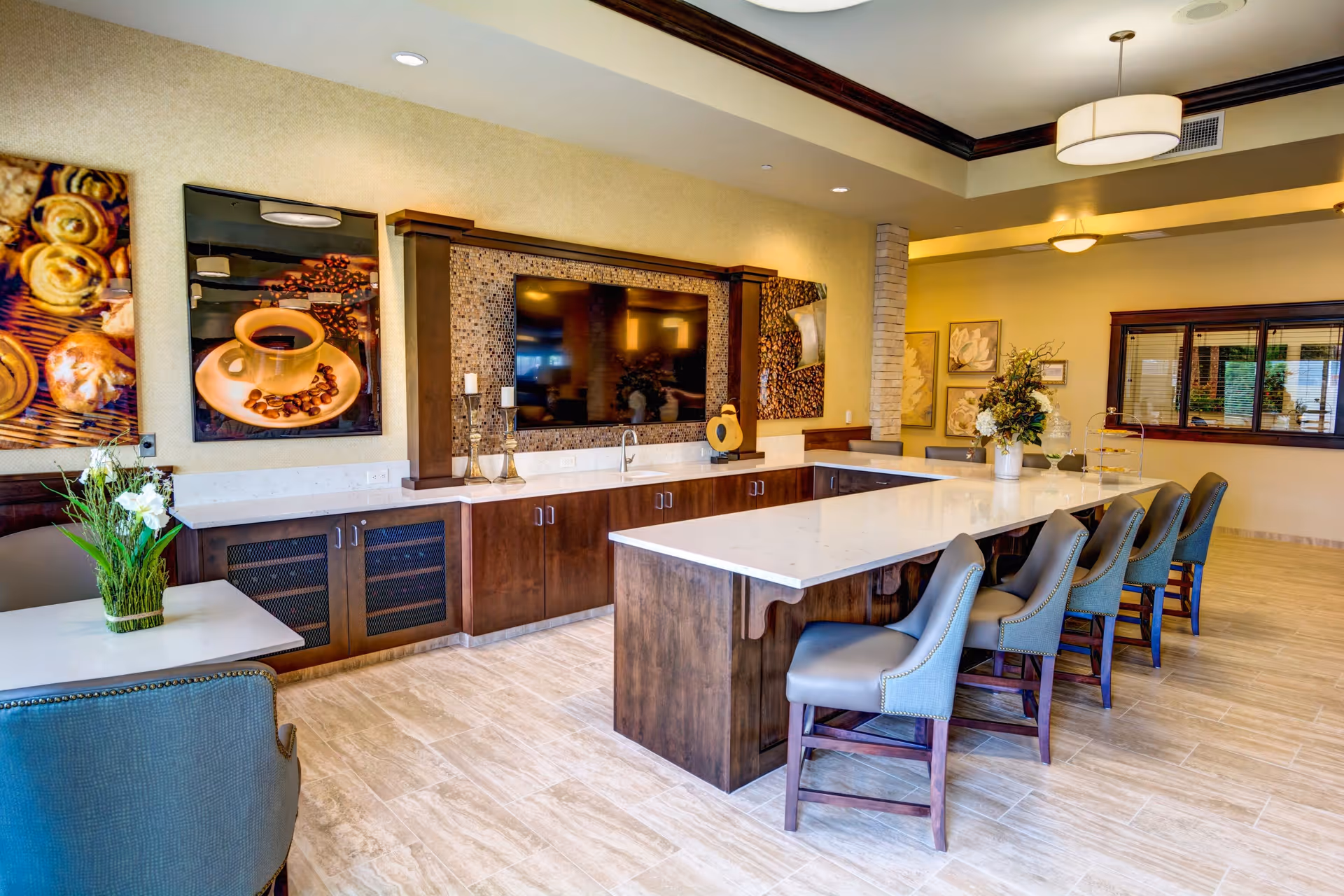A modern communal dining and lounge area with a long white countertop island surrounded by six cushioned chairs. The room features wooden cabinetry, a large flat-screen TV mounted on the wall, decorative artwork including coffee-themed pictures, and floral arrangements on the tables. The space is well-lit with ceiling lights and has a warm, inviting atmosphere.