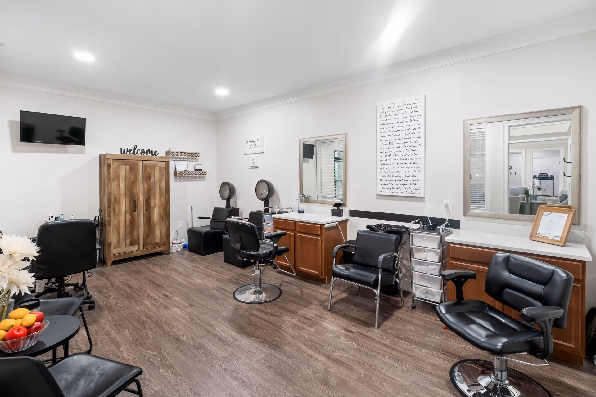Interior view of a senior living facility's salon area with multiple black salon chairs, mirrors mounted on the wall, wooden cabinets, and hair drying stations. There is a wooden cabinet with a 'welcome' sign on top, a small table with a bowl of fruit, and a wall-mounted TV. The room has wood flooring and white walls with bright ceiling lights.